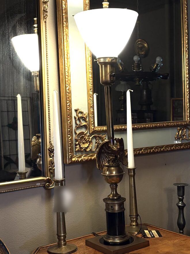 Tall brass table lamp with a cast eagle, wings spread, perched on a brass ball above a black column and square base, topped by a white glass bowl shade that throws light upward.