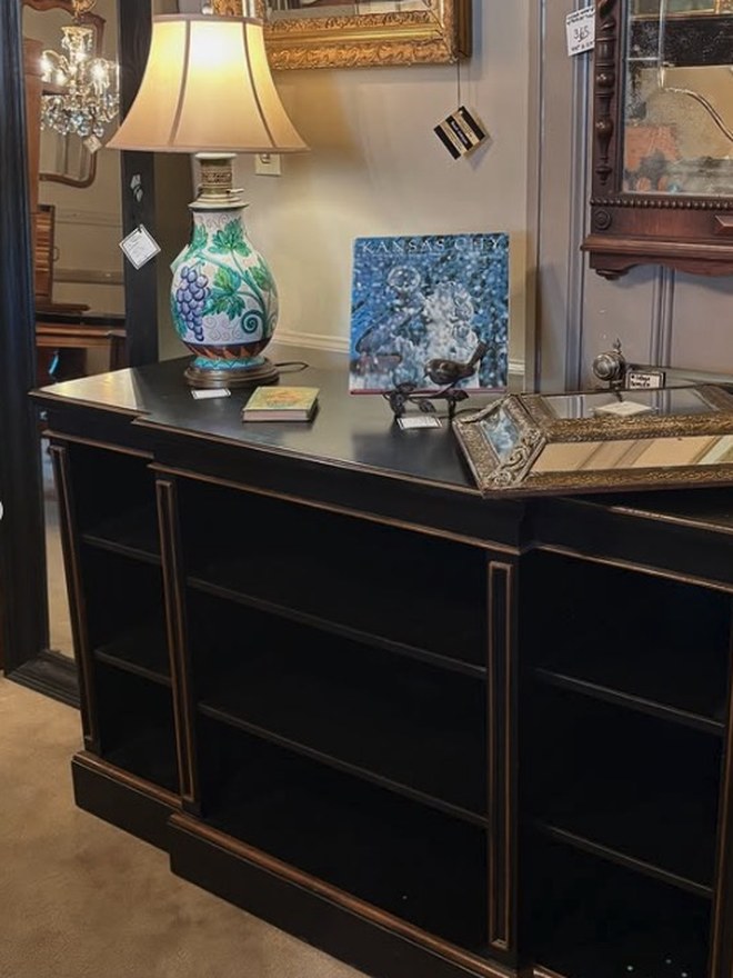 Long low ebonized open bookcase ruled with gilt lines, topped with a majolica grape-motif lamp and framed art.