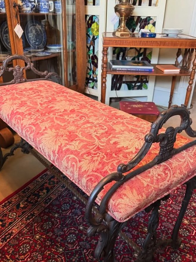 Long window bench with a scrolled cast-iron frame and rolled arms, upholstered in red-and-cream damask, on a Persian rug.