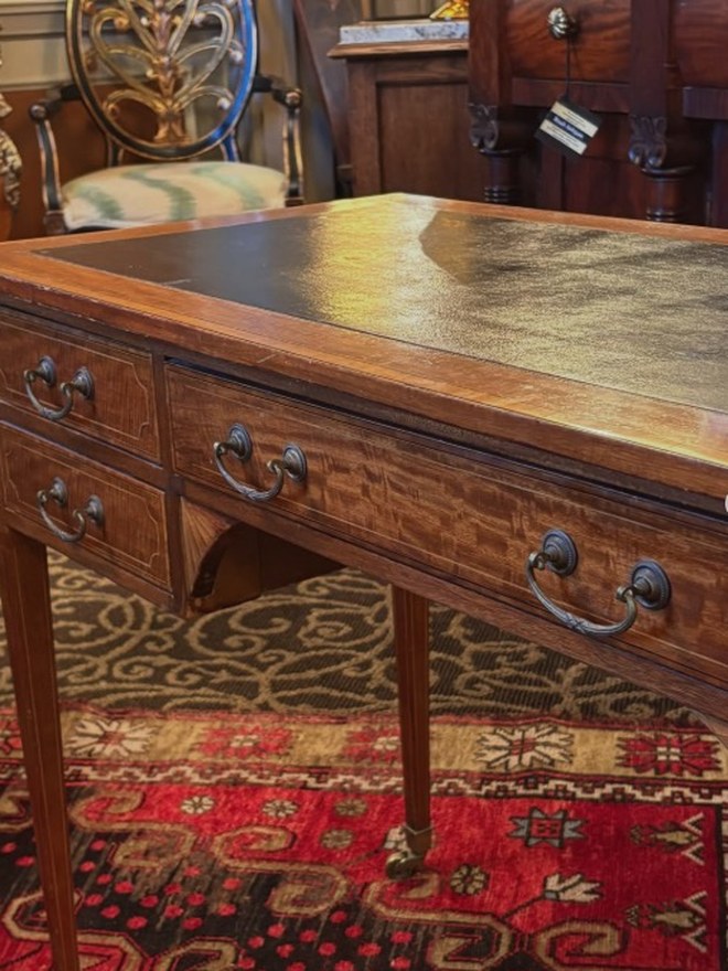 Mahogany writing table with a tooled oxblood-leather top, banded inlay, brass swan-neck handles, and brass casters, on a red rug.